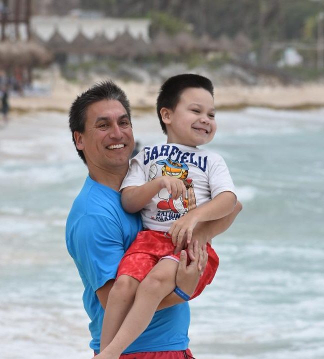 Smiling man holding smiling young boy in his arms in front of the ocean