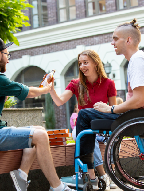 A photo with two men and one woman sitting. The man on the right is in a wheelchair and smiling. The other man is on the left, his back to the camera, and is handing the woman a juice bottle. She is in the middle facing the camera and smiling.